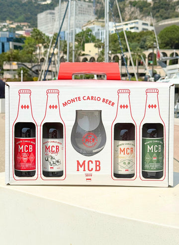 Le coffret en édition limitée avec la bière Ambrée, India Pale Ale, et les deux édititons limitées Racing et Tennis Club, accompagnés d'un verre à bière brandé Monte Carlo Beer, posé sur un banc avec en fond des bateaux et le Rocher de Monaco.