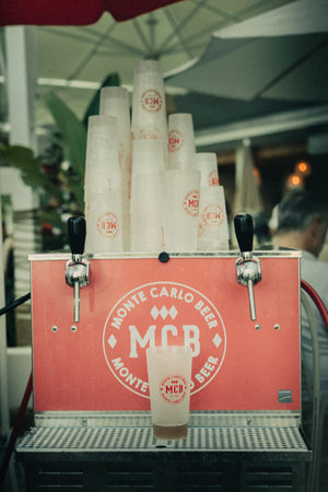 Un tireuse à bière à deux becs Monte Carlo Beer avec plusieurs Eco-cup MCB empilée et un verre à moitié remplie de bière posé sur la tireuse