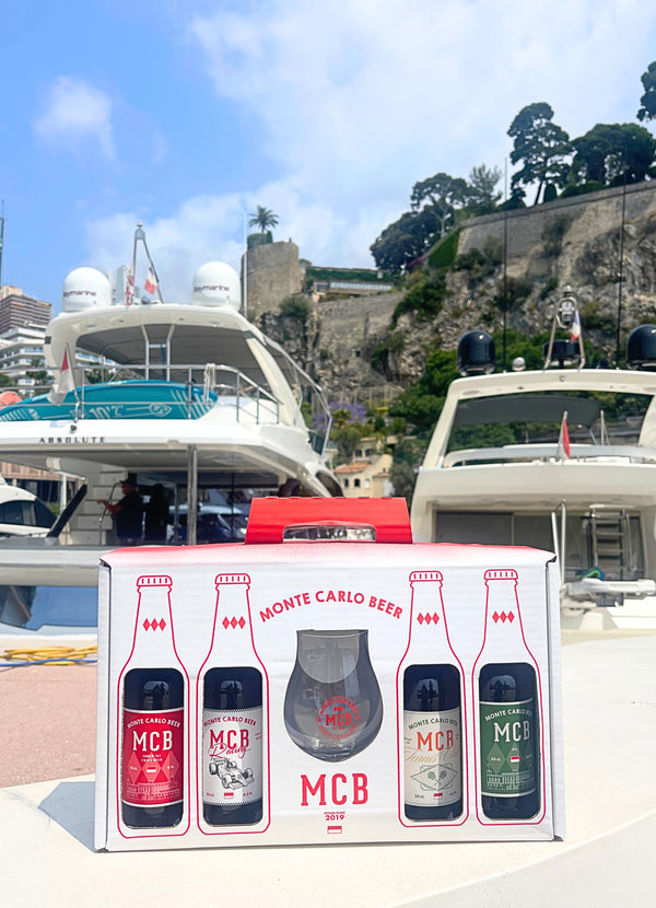 Le coffret en édition limitée avec la bière Ambrée, India Pale Ale, et les deux édititons limitées Racing et Tennis Club, accompagnés d'un verre à bière brandé Monte Carlo Beer, posé sur un banc avec en fond des bateaux et le Rocher de Monaco.